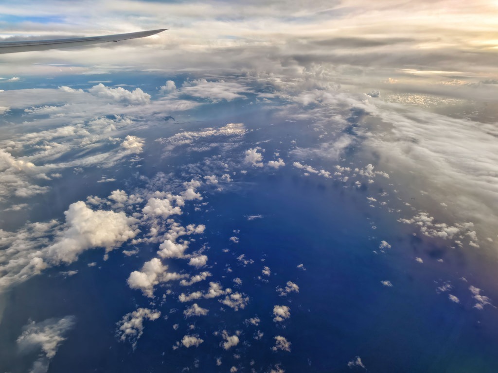 Blick aus dem Flugzeugfenster auf tiefblauen Ozean mit zahllosen kleinen weißen Wolkenfetzen, während oben links die silberne Flugzeugtragfläche und am Horizont dichtere Wolkenfelder mit sanftem Abendlicht zu sehen sind