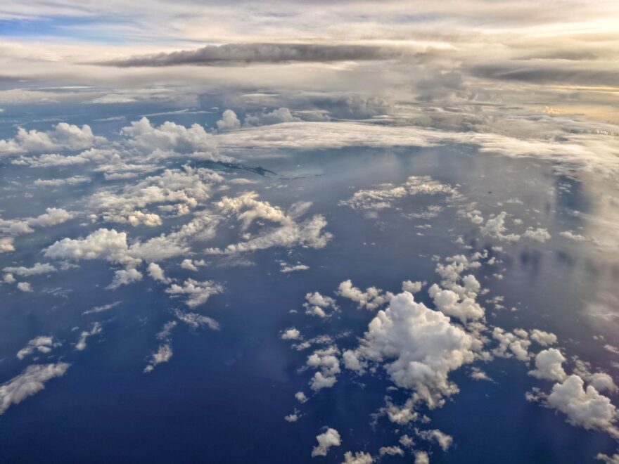Luftaufnahme eines weiten, tiefblauen Ozeans mit vereinzelten, schneeweissen Cumuluswolken im Vordergrund, während am Horizont dichte Wolkenschichten und ein sanft leuchtender Abendhimmel zu sehen sind