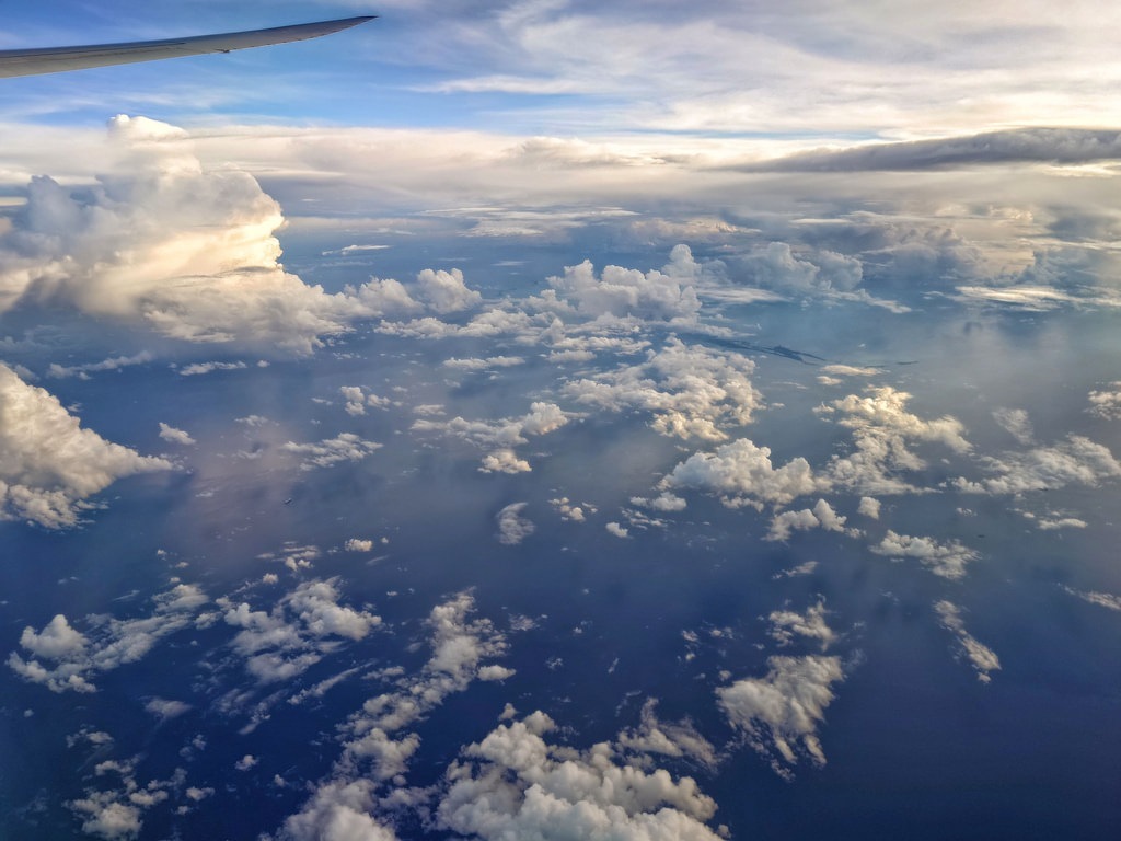 Blick aus einem Flugzeugfenster auf eine weite Wolkenlandschaft mit dichten, weissen Quellwolken, die über dem tiefblauen Ozean schweben, während am oberen Bildrand die graue Flugzeugtragfläche zu sehen ist