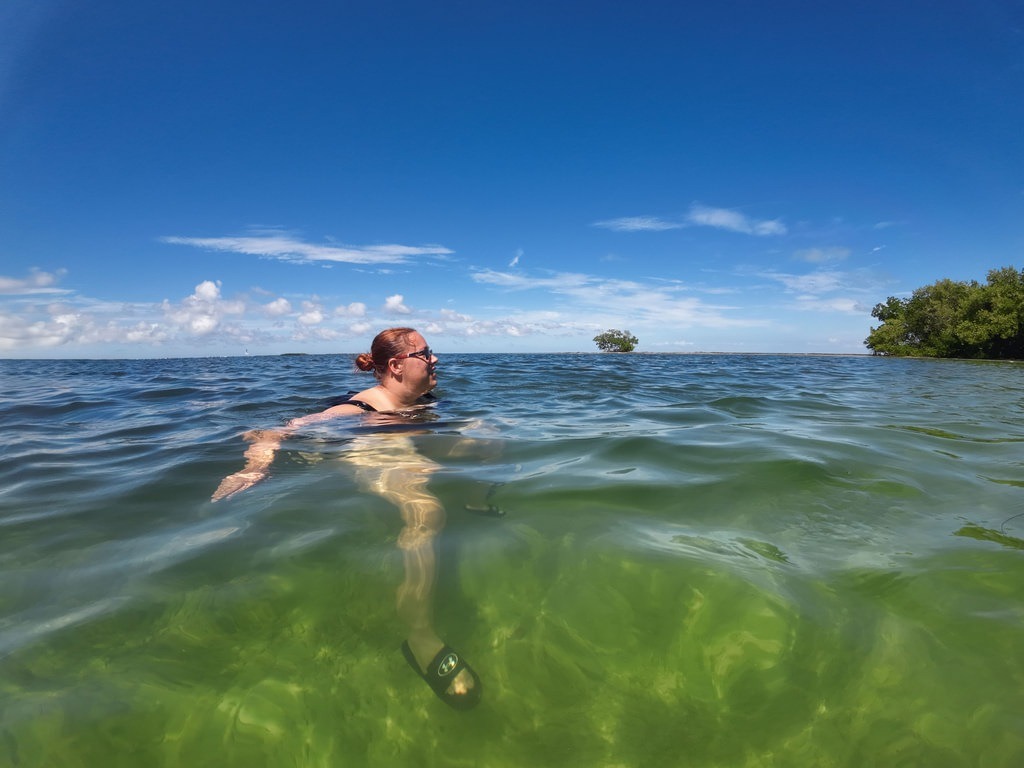 Eine rothaarige Frau mit Brille badet in grünlich-klarem Wasser vor einer Küstenlandschaft mit dichter Ufervegetation, während am blauen Himmel vereinzelte weiße Wolken ziehen